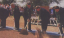 An old photograph of five men in black suits holding shovels into the dirt.